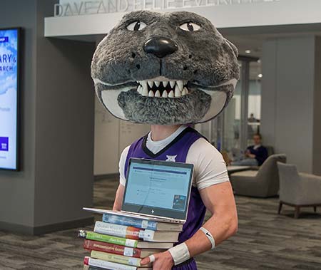 Willie Wildcat holding laptop and textbooks.